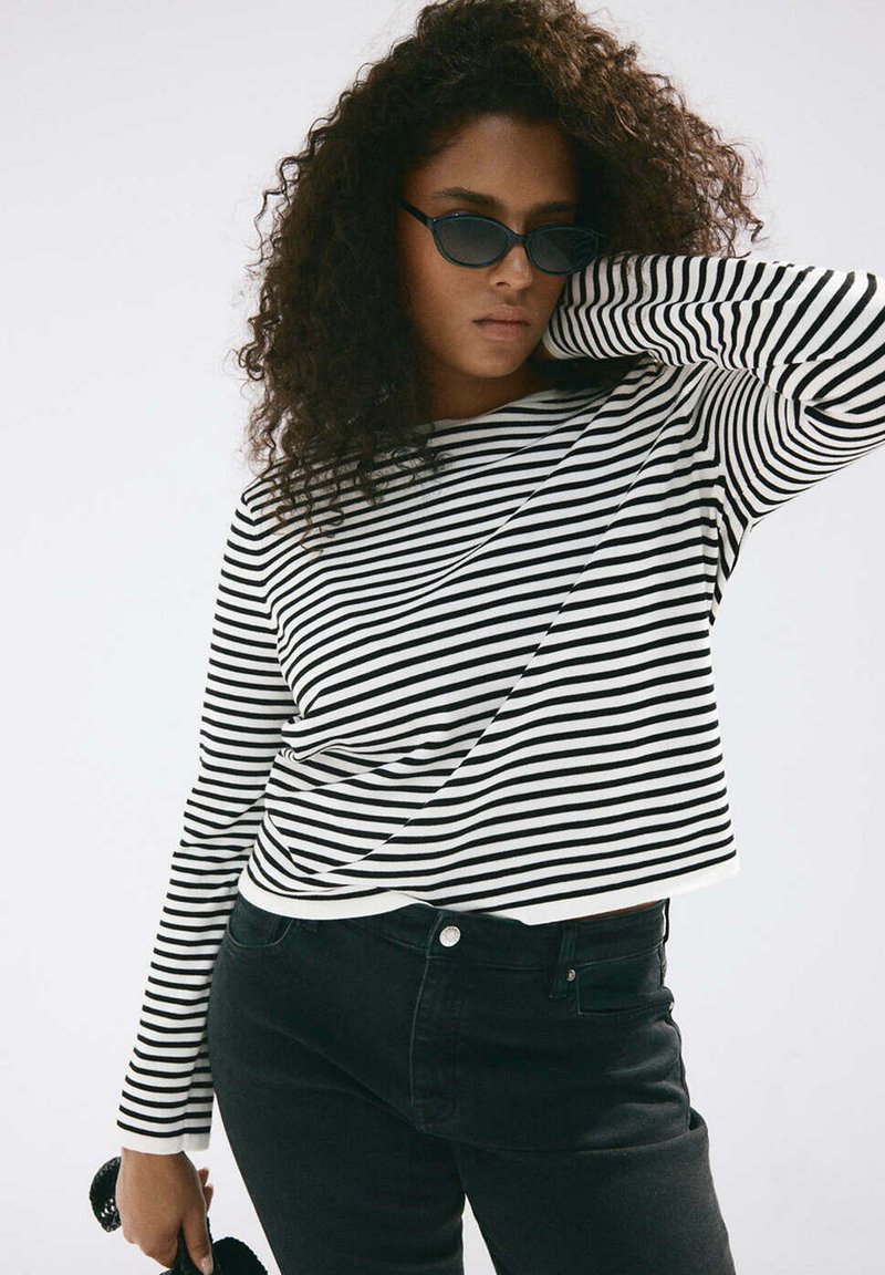 Femme aux cheveux bouclés portant des lunettes de soleil noires, un haut à manches longues rayé noir et blanc et un jean noir, posant avec un bras levé.
