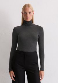 Frau mit glattem blonden Haar, die einen eng anliegenden, dunkelgrauen Rollkragenpullover und schwarze hochtaillierte Hosen trägt, steht vor einer weißen Wand.