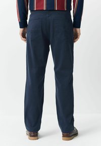 Mann mit marineblauen Hosen, braunen Lederschuhen und einem marineblauen Pullover mit vertikalen roten und weißen Streifen, der mit dem Rücken steht.