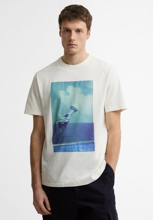 Fiatal férfi fehér pólóban, melyen kék grafika látható, kezében fagylaltkelyhet tart, háttérben az ég, sötét nadrággal.