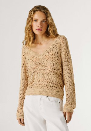 Donna con lunghi capelli biondi ricci, indossa un maglione beige a maglia aperta e pantaloni bianchi, in piedi davanti a uno sfondo neutro.