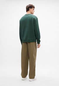 Sweatshirt verde com decote redondo, ajuste relaxado e punhos canelados, combinado com calças caqui largas e tênis brancos, visto de costas.