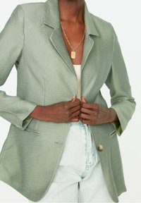 Vihreä bleiseri kevyessä kankaassa, jossa on sakara-kaulus, yksittäinen nappisulku ja kaksi etutaskua.