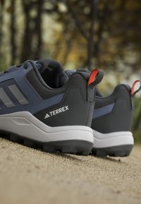 Paire de chaussures de randonnée Adidas Terrex noires et bleues reposant sur un chemin de gravier avec des arbres flous en arrière-plan.