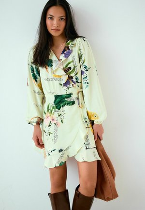 Hosszú, sötét hajú nő világos, virágos mintájú, átvetős ruhában és térdig érő barna csizmában, barna velúr táskát tartva egy fehér fal előtt.