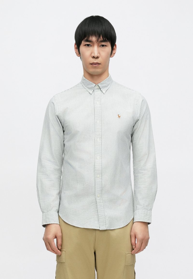 Zeleno-bílá pruhovaná košile na knoflíky s dlouhými rukávy a zaobleným lemem. Na hrudi je malé logo. Nošena s béžovými cargo kalhotami.