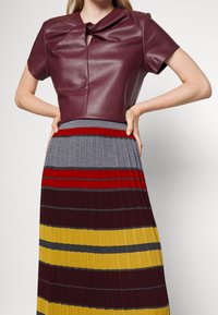 Top in finta pelle bordeaux con un dettaglio annodato al collo, abbinato a una gonna plissettata con audaci righe orizzontali in grigio, rosso e giallo.