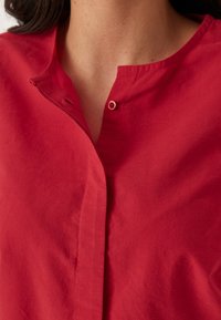 Blusa rossa in cotone con colletto alla mandarina, chiusura con un singolo bottone e tessuto leggermente strutturato. Design semplice e pulito.