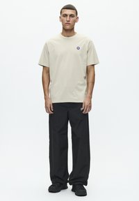 Beige bomulds t-shirt med rund hals og et lille blåt logo på venstre bryst, kombineret med løstsiddende sorte bukser og sorte sko.