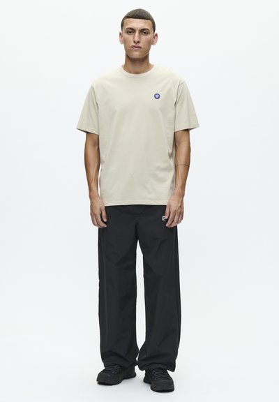 T-shirt en coton beige avec un col rond et un petit logo bleu sur la poitrine gauche, associé à un pantalon noir à coupe ample et des chaussures noires.