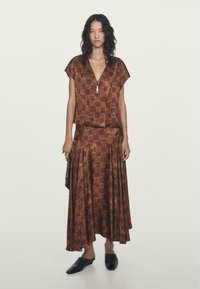 Hnědý vzorovaný dvoudílný outfit, který má top s krátkým rukávem a hlubokým výstřihem ve tvaru V a dlouhou sukni, obojí vyrobené z hladkého, splývavého materiálu.