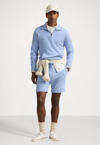 Svetlomodrý pulóver s polovičným zipsom a golierom, skombinovaný s krátkymi nohavicami rovnakej farby. Krémový sveter zviazaný v páse, pod ním biela košeľa a béžové ponožky.