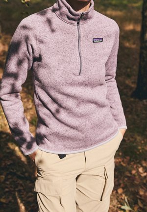 Persoon die een lichtpaarse Patagonia fleece met een kwartrits draagt en beige cargobroeken, staand buiten met handen in de zakken.