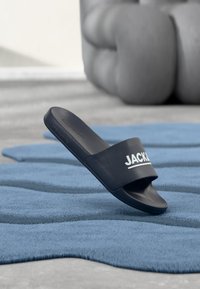 Schwarze Slidesandale mit einem breiten Riemen, der das Wort "JACK" trägt. Glattes Material, minimalistisches Design und offene Zehenpartie.