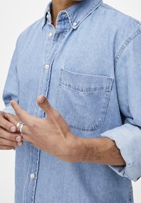 Hellblaues Jeanshemd mit einem Kentkragen, Brusttasche und sichtbaren Steppnähten. Die Hand der Person hebt durch Gesten die Ringe hervor.