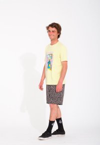 Camiseta gráfica de color amarillo claro con ilustración colorida, combinada con pantalones cortos negros con un patrón abstracto blanco. Calcetines y zapatos negros.