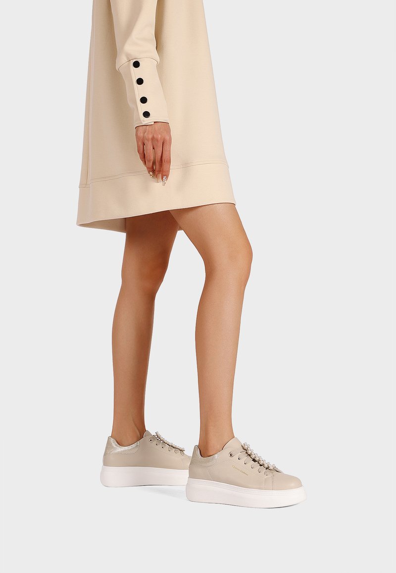 Sneakers beige con tomaia in pelle testurizzata, dettagli argento e suola piatta bianca. Esposti accanto a un vestito beige con bottoni neri.