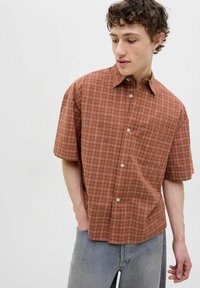Camisa de cuadros marrón de manga corta con cuello abotonado, hecha de tejido ligero; presenta botones blancos y un único bolsillo en el pecho.