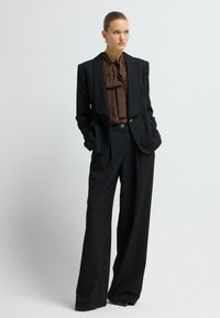 Schwarzer maßgeschneiderter Anzug mit weitem Bein, ausgestattet mit einer braunen Bluse mit Schleife und silbernen Akzentknöpfen am Blazer.