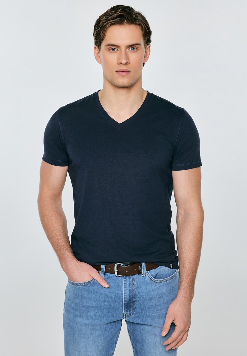 Ung mand med kort brunt hår iført en tætsiddende sort T-shirt med V-udskæring, blå jeans og en brun bælte, stående med den ene hånd i lommen.