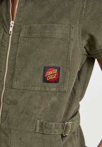 Salopette in corduroy verde oliva con zip frontale, tasca a toppa e dettaglio della cintura. Presenta un logo rosso e nero di Santa Cruz sulla tasca.