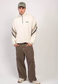 Giovane uomo con maglione beige con zip a quarto, righe multicolori sulle maniche, pantaloni cargo marroni larghi, sneakers mimetiche e cappello verde oliva, in piedi con le mani nelle tasche.