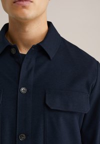 Marinblå knappad skjorta i ett texturerat tyg, med spridd krage, två bröstfickor och svarta knappar.
