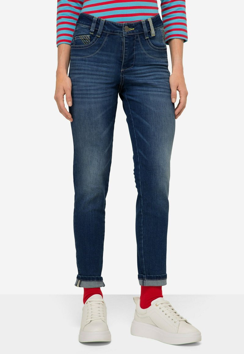 Granatowe dżinsy z średnim stanem, z podwiniętymi mankietami i kontrastowym przeszyciem. Połączone z białymi sneakersami i czerwonymi skarpetkami.