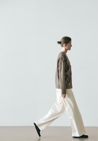 Kobieta idąca w prawo, ubrana w brązowy kardigan w romby, białe spodnie o szerokich nogawkach oraz czarne buty na jednolitym jasnoszarym tle.