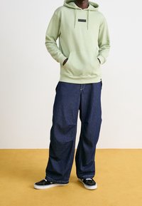 Felpa con cappuccio verde chiaro con tasca frontale e toppa con logo, abbinata a jeans larghi blu scuro e sneakers nere con suole bianche.