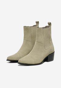 Botines de ante beige hasta el tobillo con puntera puntiaguda y tacón bajo. Presentan paneles laterales elásticos y tiradores para facilitar su puesta.