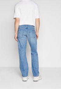 Hombre con jeans rectos de color azul claro, zapatillas blancas y una camiseta de manga corta blanca, de pie contra un fondo claro y liso.