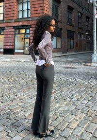 Črtasta majica z dolgimi rokavi in belim ovratnikom, v kombinaciji z oprijetimi, širokimi sivo-črnimi hlačami. Manekenka nosi črne čevlje in stoji na tlaku iz kock.