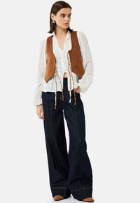 Gilet di camoscio marrone sopra una camicia bianca con bottoni e texture, abbinato a jeans in denim scuro a gamba larga e stivali marroni con punta a punta.
