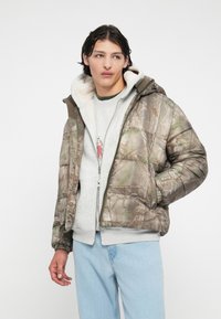 Kamutömbszerű kabát álcázó mintázattal, szőrme bélésű kapucnival, barna és zöld árnyalatokkal, cipzáras záródással és rugalmas mandzsettákkal, szürke pulóver és világos farmernadrág fölött.