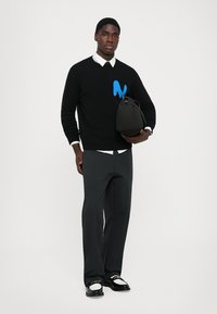 Pulover negru tricotat cu litera albastră "M", asortat cu pantaloni gri și pantofi negri. Bărbatul ține o geantă mică neagră. Fundal neutru.