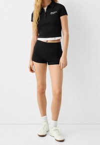 Femme portant un polo noir court avec le texte "59th Street New York", un short noir, des chaussettes blanches et des baskets blanches, se tenant contre un fond blanc.