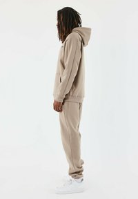 Conjunto de sudadera beige y pantalones de chándal, con un corte relajado, bolsillo canguro y puños acanalados, combinado con zapatillas blancas.