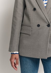 Blazer doppiopetto in motivo scozzese, grigio e nero. Presenta grandi bottoni neri, tasche laterali e un tessuto texture. La manica con risvolto mostra uno strato in maglia con accenti blu sotto.