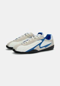 Beige, fehér és kék sportcipők, sima bőr felsőrésszel, párnázott bokarészsel és texturált tapadású fekete gumitalppal.