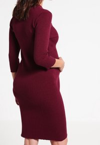 Robe bodycon tricotée bordeaux avec texture côtelée, manches trois-quarts et ourlet mi-genou, offrant une coupe ajustée au niveau du torse.