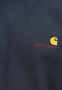 Φούτερ σε ναυτικό μπλε με ριμπ λαιμόκοψη και διακοσμητική ραφή σε σχήμα V. Διαθέτει κόκκινο κείμενο "carhartt" και κίτρινο λογότυπο στην αριστερή πλευρά του στήθους.