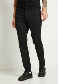 Pantaloni neri dal design slim fit, realizzati in tessuto liscio. Presentano un frontale piatto, tasche laterali e sono indossati con sneakers nere.