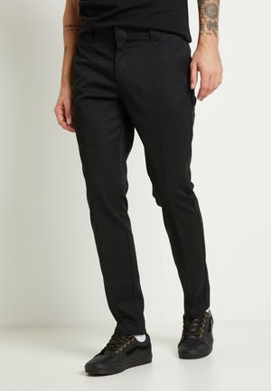 Pantalones negros de corte ajustado, hechos de una tela suave. Con frentes planos, bolsillos laterales y combinados con zapatillas negras.