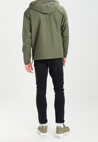 Lättviktsjacka i olivgrönt med huva, svarta tajta byxor och olivgröna sneakers med vita sulor. Enkel design utan synliga mönster.