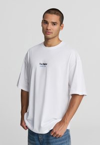 T-shirt bianco in cotone con vestibilità oversize, caratterizzato da testo ricamato blu "Los Angeles" sul davanti. Il modello indossa jeans azzurri.