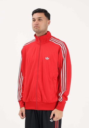 Giovane uomo che indossa una giacca da ginnastica rossa Adidas con strisce bianche e pantaloni neri, in piedi davanti a uno sfondo bianco semplice.