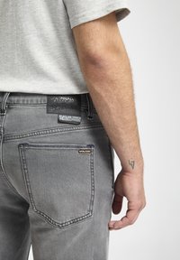 Gray denim jeans with a black leather brand patch on the back and a small orange logo. Features a classic five-pocket design and detailed stitching.