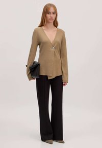 Beige wikkeltrui met een diepe V-hals en zilveren knoopdetails, gecombineerd met een hoge, zwarte broek en puntige beige hakken. Zorgt voor een moderne uitstraling.