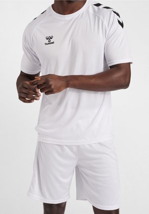 Personne à la peau foncée portant un maillot et un short de sport blanc de la marque Hummel, les mains jointes devant, debout devant un fond uni.